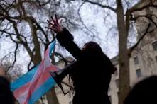 Eine Person hält ein Megafon, Im Hintergrund Bäume und Häuser, links neben der Person weht eine trans Flagge
